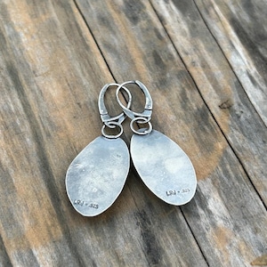 May include: A pair of silver earrings with a teardrop shape. The earrings have a silver hook closure and are marked "LRJ 925" on the back.