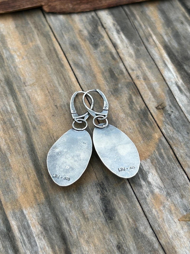May include: A pair of silver earrings with a teardrop shape. The earrings have a silver hook closure and are marked "LRJ 925" on the back.