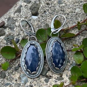 May include: A pair of silver earrings with a dark blue gemstone set in a silver oval frame. The earrings have a simple hook closure.