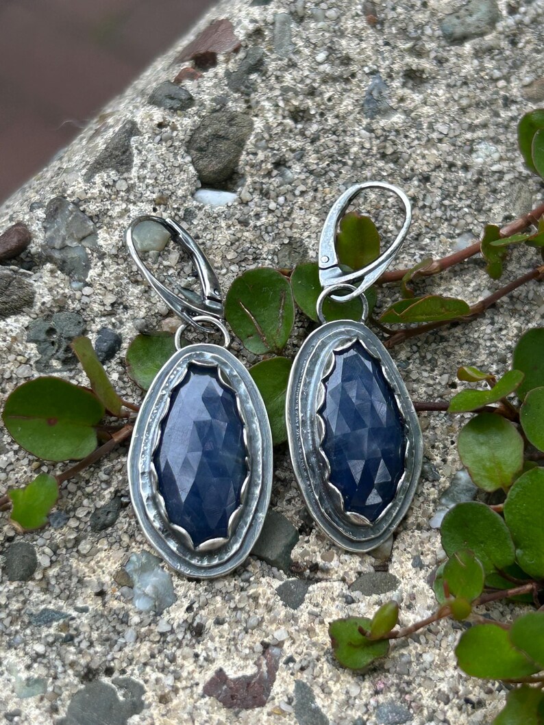 May include: A pair of silver earrings with a dark blue gemstone set in a silver oval frame. The earrings have a simple hook closure.