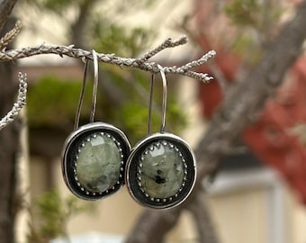 May include: A pair of silver drop earrings with oval, light green gemstones. The gemstones are set in a silver bezel with a beaded edge. The earrings are hanging from a tree branch against a blurred background.