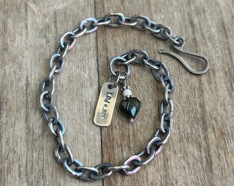 Puede incluir: Pulsera de cadena de plata con cierre de gancho. Una pequeña etiqueta de latón con texto y una cuenta verde facetada cuelgan de la cadena. La pulsera se muestra sobre una superficie de madera.