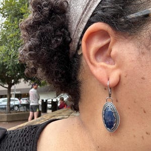 May include: A pair of silver earrings with a dark blue gemstone set in a silver bezel. The earrings are dangling from a silver hook.
