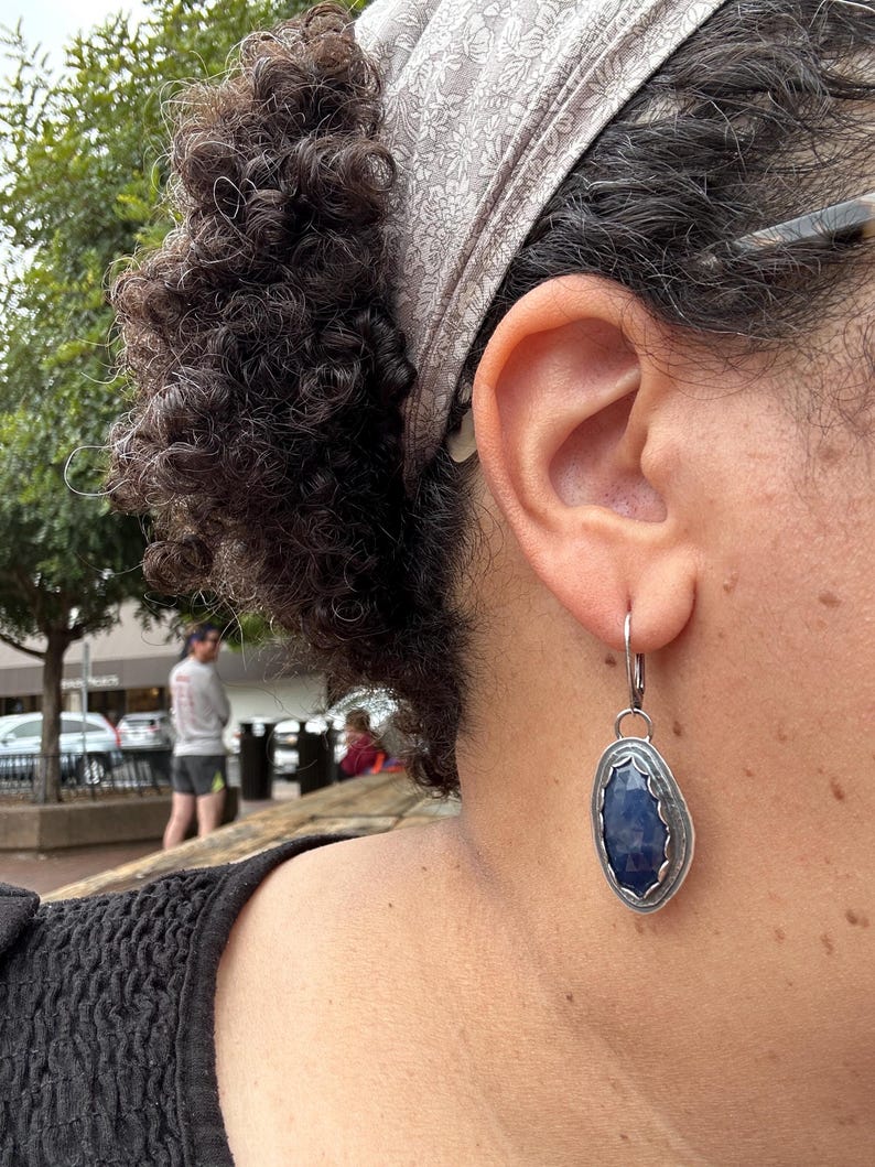 May include: A pair of silver earrings with a dark blue gemstone set in a silver bezel. The earrings are dangling from a silver hook.