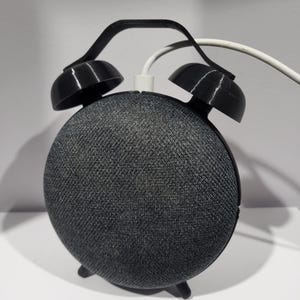 May include: A black and gray smart speaker with a custom alarm clock design. The speaker has a fabric-covered body and black plastic alarm clock features, including bells and a handle. A white cable is plugged into the speaker.