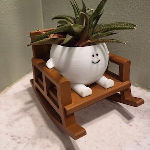 May include: A white, spherical planter with a simple black smiley face graphic, featuring small white arms and legs, seated in a brown, 3D-printed rocking chair. A green, spiky succulent plant is potted inside the white, round container.