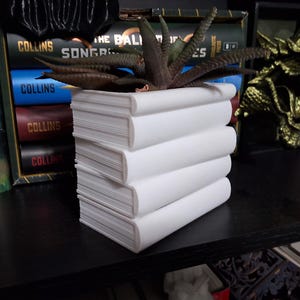 May include: A white ceramic planter shaped like a stack of books, holding a succulent plant. The planter has a clean, minimalist design, with the book spines facing outward. The plant has dark green and brown leaves. The planter is on a black shelf.