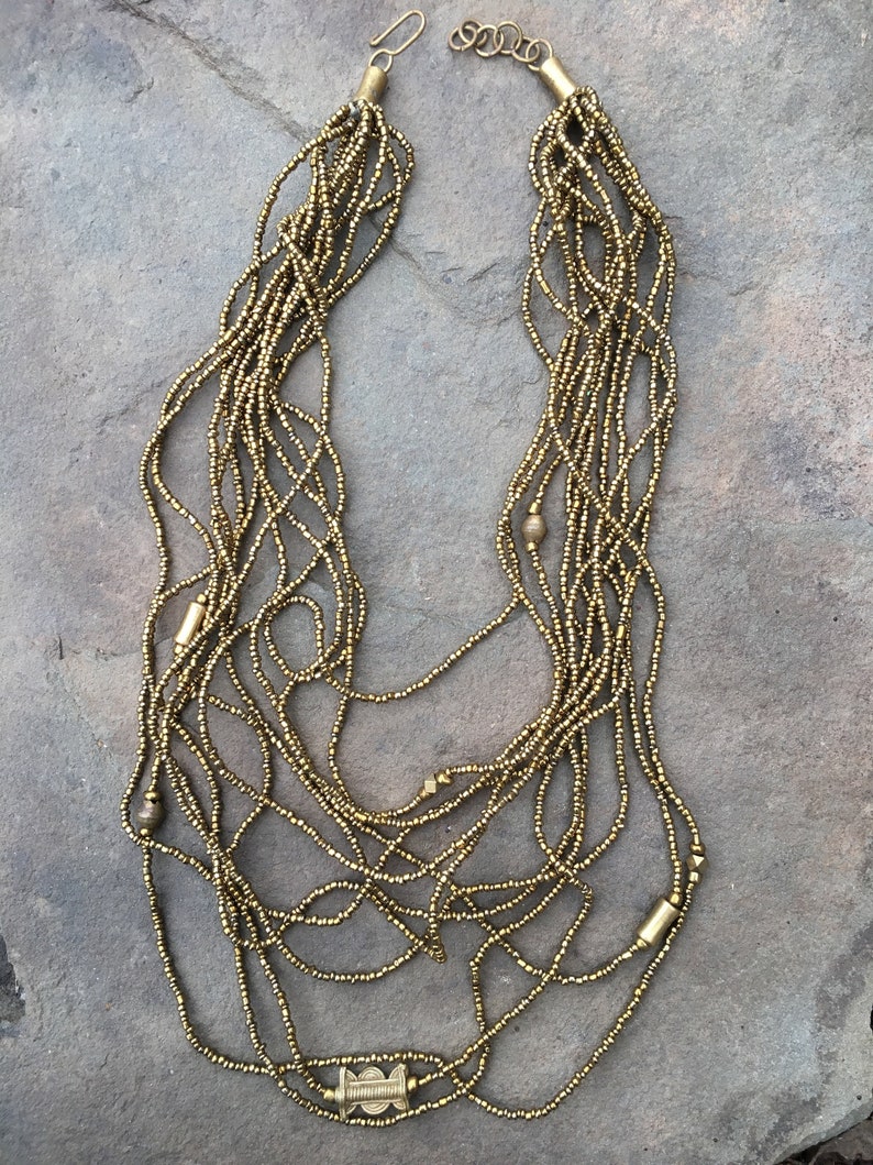 K&ouml;nnte beinhalten: Eine goldene Perlenkette mit mehreren Str&auml;ngen und einem dekorativen Verschluss. Die Kette besteht aus kleinen, runden Perlen und hat ein schlichtes, elegantes Design.