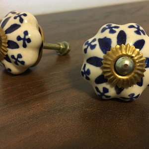 May include: Two white ceramic cabinet knobs with blue floral patterns. One knob has a gold-colored metal base with a silver center. The other knob has a silver metal base.