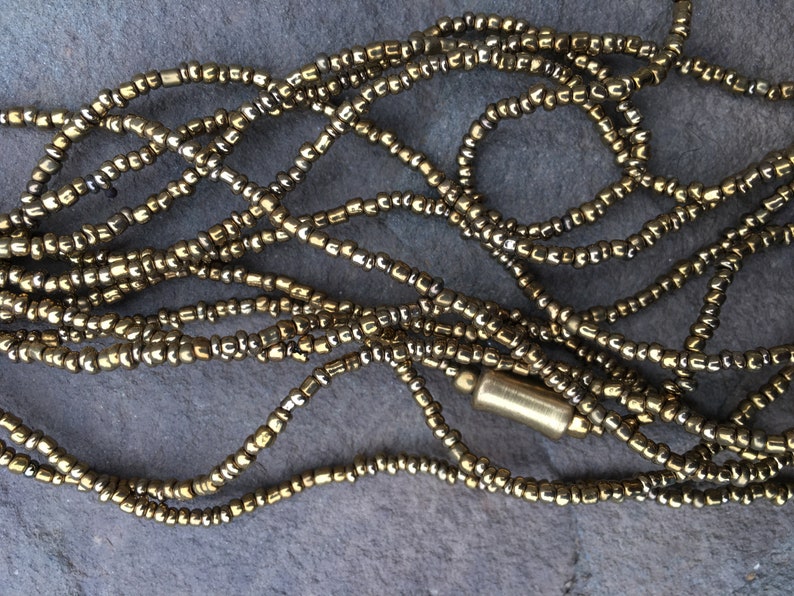 K&ouml;nnte beinhalten: Eine goldene Perlenkette mit einem goldenen Metallverschluss. Die Kette besteht aus kleinen, runden Perlen, die zu einer langen Schnur zusammengef&uuml;gt sind.