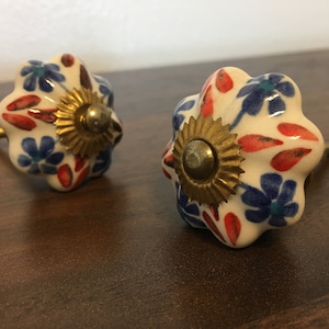 May include: Two ceramic cabinet knobs with a white base and a floral design in red, blue, and green. The knobs have a gold metal base with a screw.