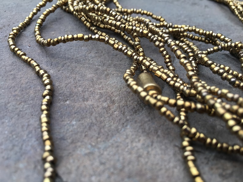K&ouml;nnte beinhalten: Eine goldene Perlenkette mit einem goldenen Metallverschluss. Die Kette besteht aus mehreren Perlenstr&auml;ngen und liegt auf einer grauen Oberfl&auml;che.