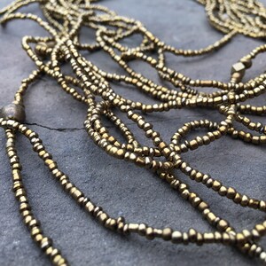 K&ouml;nnte beinhalten: Eine goldene Perlenkette mit mehreren Str&auml;ngen, an einem Ende mit einer gro&szlig;en runden Perle und am anderen Ende mit einer quadratischen Perle.