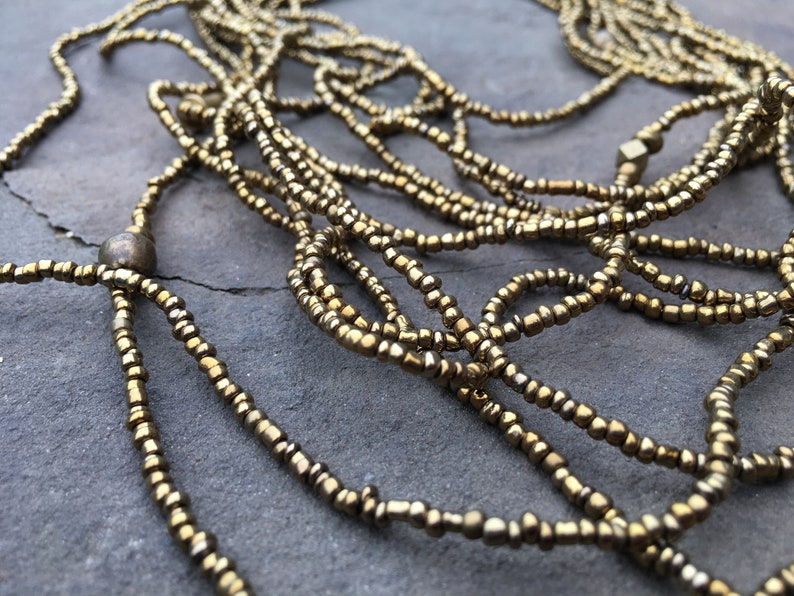 K&ouml;nnte beinhalten: Eine goldene Perlenkette mit mehreren Str&auml;ngen, an einem Ende mit einer gro&szlig;en runden Perle und am anderen Ende mit einer quadratischen Perle.