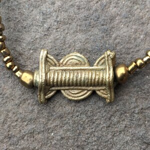 K&ouml;nnte beinhalten: Ein goldfarbenes Perlenarmband mit einem dekorativen goldenen Anh&auml;nger in der Mitte. Der Anh&auml;nger hat ein kreisf&ouml;rmiges Design mit einer erhabenen Mitte und einer strukturierten Oberfl&auml;che.