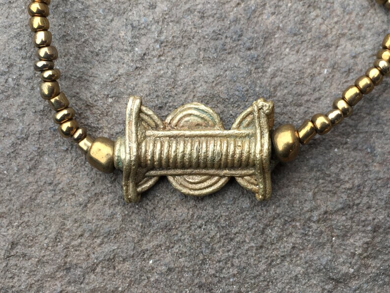 K&ouml;nnte beinhalten: Ein goldfarbenes Perlenarmband mit einem dekorativen goldenen Anh&auml;nger in der Mitte. Der Anh&auml;nger hat ein kreisf&ouml;rmiges Design mit einer erhabenen Mitte und einer strukturierten Oberfl&auml;che.