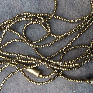 K&ouml;nnte beinhalten: Eine Nahaufnahme einer goldenen und silbernen Perlenkette. Die Kette besteht aus kleinen, runden Perlen, die zu einer langen Schnur zusammengef&uuml;gt sind. Die Perlen sind in einem zuf&auml;lligen Muster angeordnet, was ein einzigartiges und auff&auml;lliges Design ergibt.