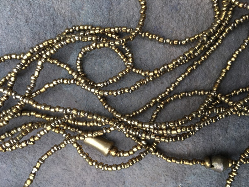 K&ouml;nnte beinhalten: Eine Nahaufnahme einer goldenen und silbernen Perlenkette. Die Kette besteht aus kleinen, runden Perlen, die zu einer langen Schnur zusammengef&uuml;gt sind. Die Perlen sind in einem zuf&auml;lligen Muster angeordnet, was ein einzigartiges und auff&auml;lliges Design ergibt.