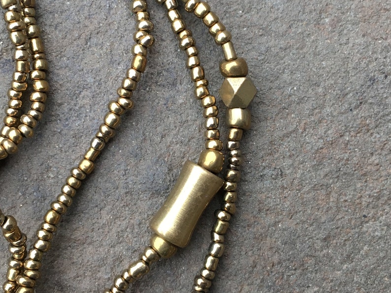 K&ouml;nnte beinhalten: Eine goldene Perlenkette mit einer zylindrischen und einer sechseckigen Perle. Die Kette besteht aus kleinen, runden Perlen.
