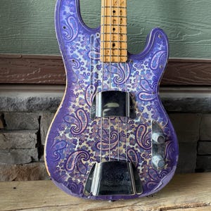 K&ouml;nnte beinhalten: Eine lila E-Bassgitarre mit einem Paisley-Muster in Blau- und Wei&szlig;t&ouml;nen. Der Bass hat einen Holz-Hals und ein Griffbrett sowie Chrom-Hardware. Der Korpus der Gitarre liegt auf einer Holzoberfl&auml;che.