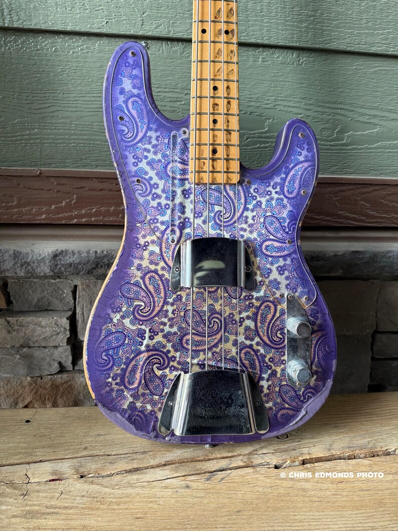 K&ouml;nnte beinhalten: Eine lila E-Bassgitarre mit einem Paisley-Muster in Blau- und Wei&szlig;t&ouml;nen. Der Bass hat einen Holz-Hals und ein Griffbrett sowie Chrom-Hardware. Der Korpus der Gitarre liegt auf einer Holzoberfl&auml;che.