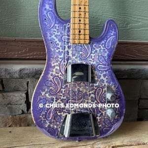 K&ouml;nnte beinhalten: Eine lila E-Bassgitarre mit Paisley-Muster. Der Korpus der Gitarre ist lila mit einem wei&szlig;en und blauen Paisley-Muster. Sie hat Chrom-Hardware und einen Holz-Hals. Der Text "CHRIS EDMONDS PHOTO" ist sichtbar.