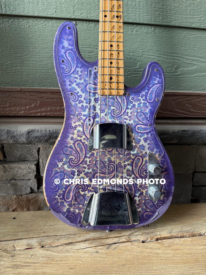 K&ouml;nnte beinhalten: Eine lila E-Bassgitarre mit Paisley-Muster. Der Korpus der Gitarre ist lila mit einem wei&szlig;en und blauen Paisley-Muster. Sie hat Chrom-Hardware und einen Holz-Hals. Der Text "CHRIS EDMONDS PHOTO" ist sichtbar.