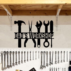 May include: A black metal wall sign featuring silhouettes of a hammer, pliers, adjustable wrench, open-end wrench, and screwdriver. The text DAD'S WORKSHOP is cut out in a bold, sans-serif font across the center of the tool silhouettes.