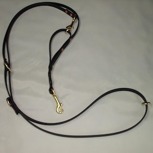 May include: A dark brown leather lead featuring multiple gold-toned metal rings and a swivel snap hook. The adjustable strap design includes visible rivets and a secondary attachment point, creating a versatile configuration for pet handling.