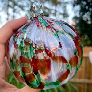May include: A hand-blown glass ornament with a clear loop for hanging. The ornament is a round shape with a ribbed texture. It features a mix of colors, including green, red, and white, creating a unique, decorative piece.