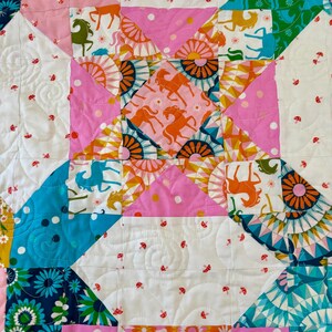 Puede incluir: Una colcha de patchwork con un patr&oacute;n geom&eacute;trico repetitivo de cuadrados y tri&aacute;ngulos. La tela muestra una variedad de estampados coloridos que incluyen caballos estilizados, motivos florales de sol y peque&ntilde;os hongos rojos sobre un fondo blanco.
