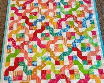 Puede incluir: Una colcha de retazos colorida con un fondo blanco y una variedad de colores brillantes, incluyendo rosa, rojo, verde, azul y amarillo. La colcha está hecha de cuadrados y rectángulos, y el patrón es un poco abstracto. La colcha está bordeada con un ribete azul claro.