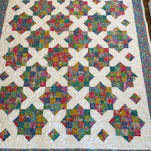 May include: A white quilt with a blue and green floral border. The quilt is made up of a patchwork of colorful squares, arranged in a star pattern.