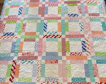 Puede incluir: Un edredón de retazos colorido con un fondo blanco y cuadrados de varias telas estampadas en rojo, azul, verde y naranja. El edredón tiene un borde de lunares azul claro y acolchado floral.