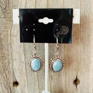 May include: A pair of silver-toned earrings with a light blue oval stone. The earrings have a decorative scalloped edge around the stone and a hook closure. The earrings are displayed on a white stand against a wooden background.