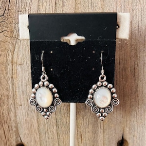 May include: A pair of silver-toned earrings with a white oval center stone. The earrings have a decorative border of small beads and swirling filigree. The earrings are displayed on a black card against a wooden background.