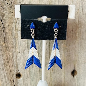 May include: A pair of blue and white arrow-shaped earrings. The earrings have a blue enamel finish with white accents. They are displayed on a black earring card against a wooden background.