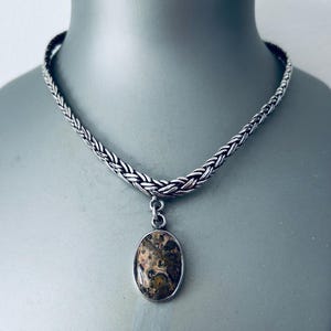 May include: A silver-toned necklace with a braided chain and an oval pendant. The pendant features a speckled pattern in shades of brown, tan, and green, set in a silver frame. The necklace is displayed on a mannequin.