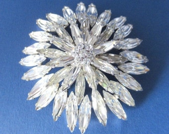 Puede incluir: Un broche grande de tono plateado con un diseño de flor. El broche está hecho de muchas pequeñas piedras brillantes transparentes que están dispuestas en un patrón circular. El centro del broche es un grupo de piedras brillantes más pequeñas.