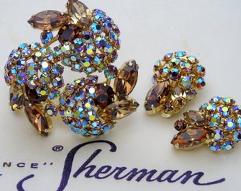 May include: A vintage brooch and earrings set featuring a cluster of iridescent rhinestones and brown faceted stones. The set is displayed on a white background with the text "Sherman" in purple.