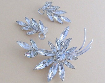 Puede incluir: Un conjunto de joyas de pedrería vintage que incluye un broche grande con diseño floral y dos pendientes más pequeños con diseños de hojas. Las joyas están hechas de metal plateado y pedrería transparente.