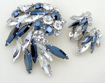 Puede incluir: Un conjunto vintage de broche y pendientes con un grupo de piedras de cristal transparentes y azul oscuro. El broche tiene forma de abanico y los pendientes son de clip.