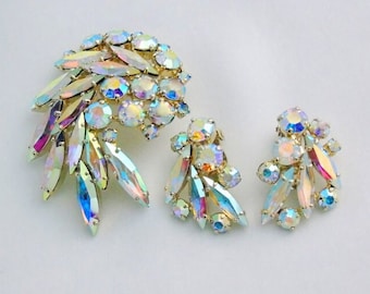 Puede incluir: Un conjunto de joyas de cristal iridiscente que incluye un broche y un par de pendientes. El broche tiene forma de racimo de hojas con un cristal grande en el centro. Los pendientes también tienen forma de hojas con un cristal más pequeño en el centro.