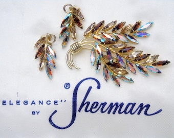 May include: A gold tone brooch and earrings set with iridescent brown and blue rhinestones. The brooch is shaped like a branch with leaves. The set is labeled "Elegance' by Sherman".