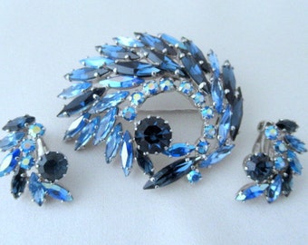 May include: A vintage blue rhinestone brooch and earrings set. The brooch features a swirl design with a large blue rhinestone in the center. The earrings are smaller and have a similar design.