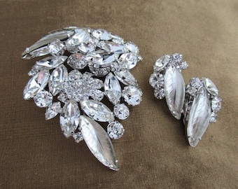 May include: A silver tone brooch and earrings set with clear rhinestones. The brooch is a cluster of large and small rhinestones in a leaf shape. The earrings are also leaf shaped with rhinestones.