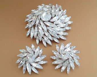 Puede incluir: Un conjunto de tres piezas de joyería de diamantes de imitación brillantes, que incluye un broche grande y dos pendientes a juego. El broche es un diseño grande y circular con un diamante de imitación central y filas radiantes de diamantes de imitación más pequeños. Los pendientes son diseños circulares más pequeños con un diamante de imitación central y filas radiantes de diamantes de imitación más pequeños.