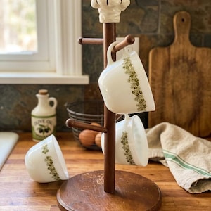 Puede incluir: Un árbol de tazas de madera con una gorra de chef en la parte superior sostiene tres tazas blancas con diseños florales verdes. Una taza descansa sobre la encimera. El árbol está sobre una superficie de madera, con elementos de cocina y una ventana al fondo.