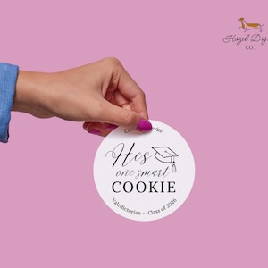以下が含まれることがあります： 「He's one smart COOKIE」の文字と卒業帽子のイラストが描かれた白い丸いステッカー。また、「Valedictorian - Class of 2026」も含まれています。ステッカーはピンク色の背景に保持されています。