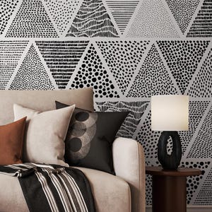 May include: A living room scene featuring a beige sofa with several throw pillows in shades of brown and black. A black and white patterned throw blanket is draped over the sofa. The wall behind the sofa is decorated with a black and white triangle pattern.